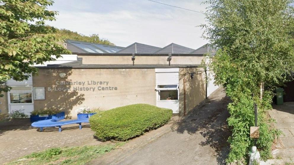 A single-storey, cream-coloured stone building, with a paved area to the front where there is a blue 'S'-shaped bench.