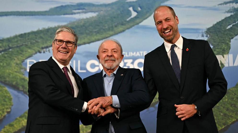 From left to right, Sir Keir Starmer, President Lula and Prince William. All are wearing dark-coloured suits. Sir Keir is wearing a white shirt and dark red tie; President Lula a light blue shirt; and Prince William a white shirt and blue tie. They are putting their hands together in an apparent show of unity. Blue and green COP30 banners are in the background.