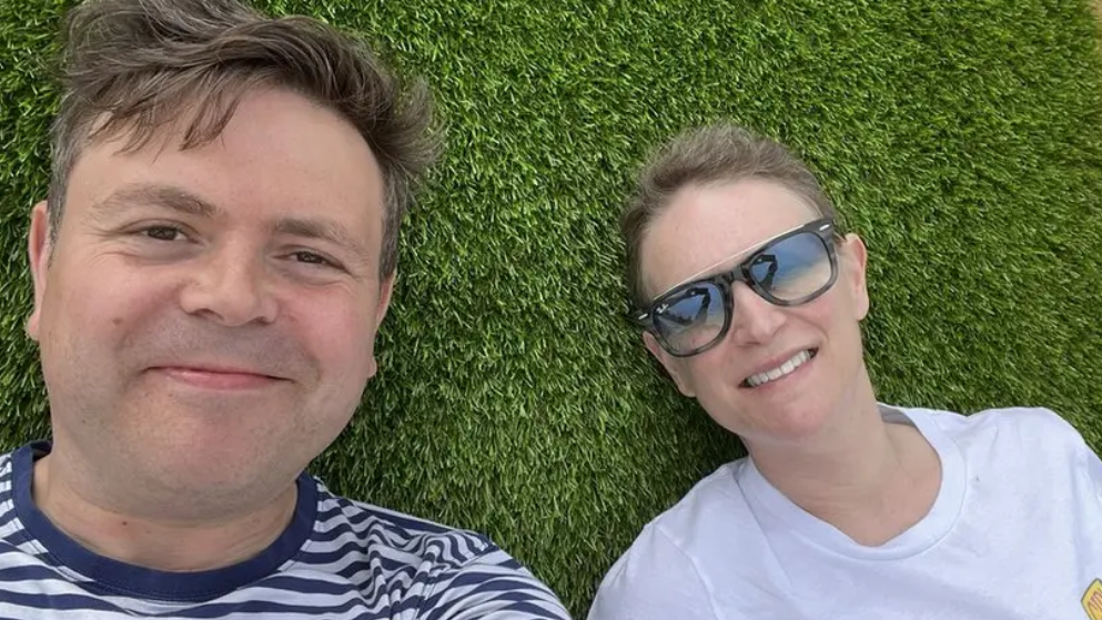 Geraint and Deb lying on grass looking into the camera. He is wearing a navy and white striped t-shit and has short brown hair. She has her blonde hair tied back and is wearing sunglasses and a white t-shirt.