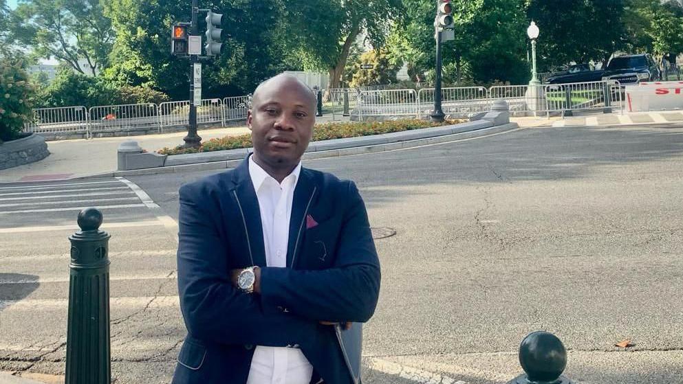 Dr Yves Kaduli, a 38-year-old asylum seeker from the Democratic Republic of Congo, stands in front of the Capitol Building with his arms folded in a suit jacket 