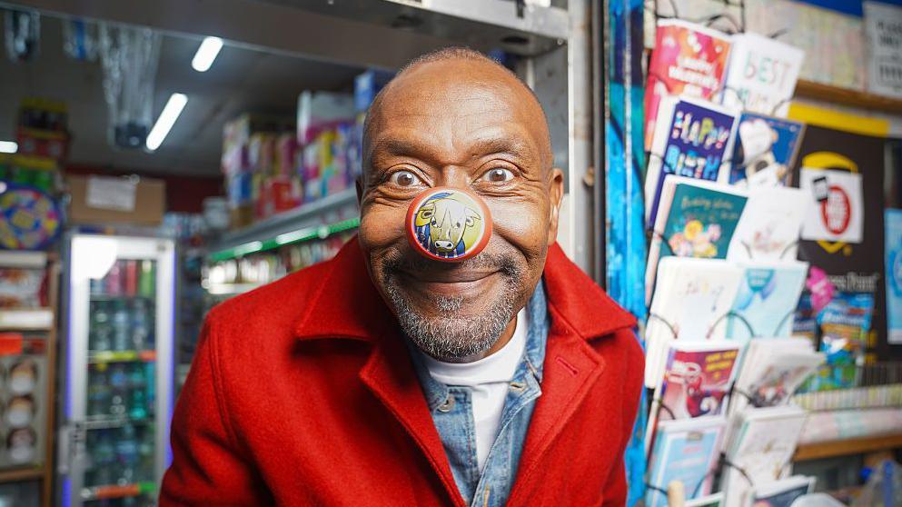 sir lenny henry wearing a red coat and a red nose standing outside a corner shop.
