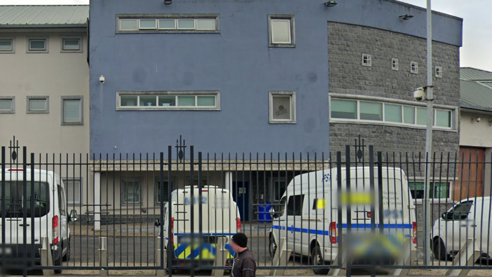 A white wall prison with three stories and blue front. Six vehicles are parked outside, including a police car. 