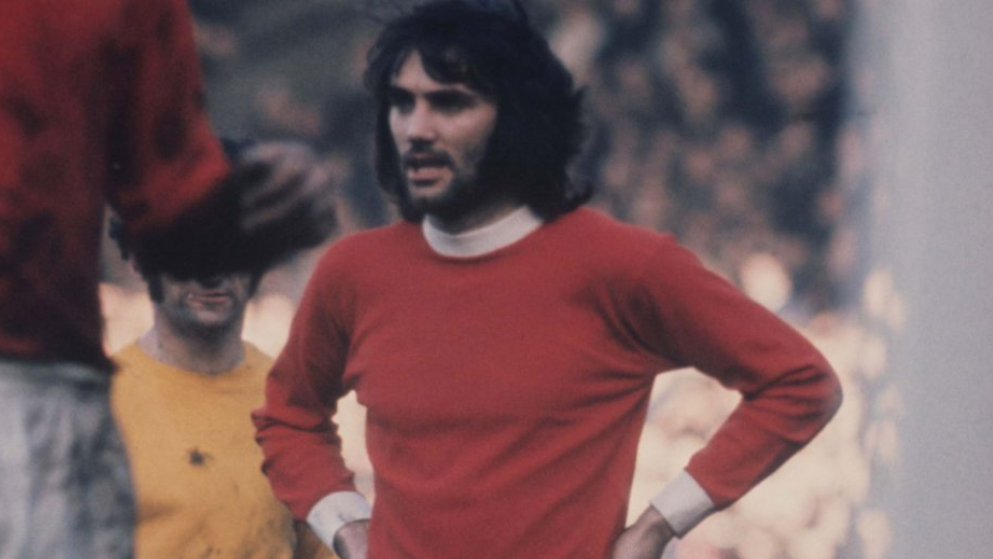 Dark-haired George Best, with a thick black beard, has his hands on his hips while wearing a red Manchester United shirt with white collar and cuffs