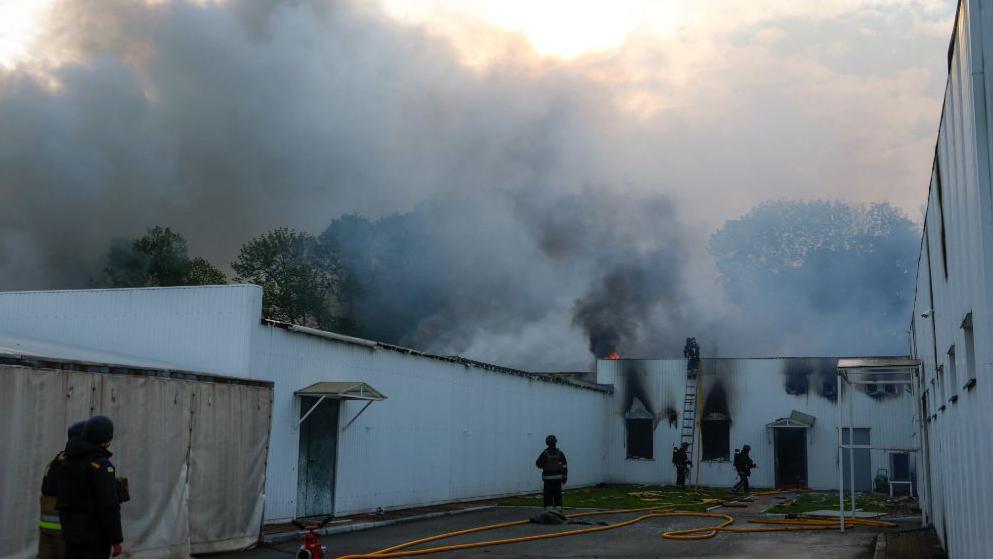 Firefighters at a site outside Kyiv scale a blackened wall as a massive smoke cloud looms ahead of them