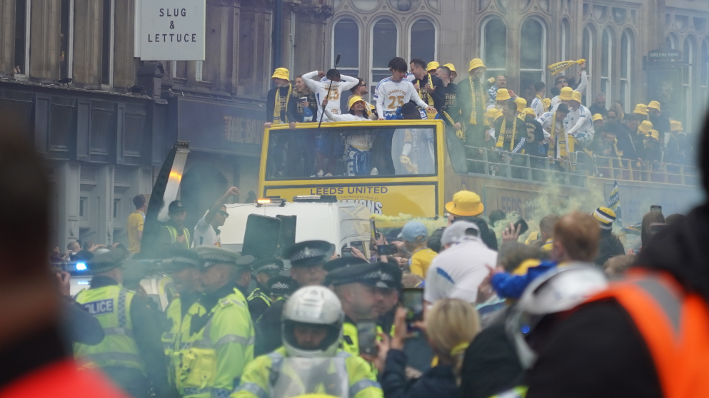 A yellow open top bus moves through crowds in Leeds. Leeds United football players are on top of the bus wearing yellow bucket hats and scarves. Police and vans escort on the street below.