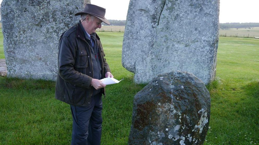 Side profile of Richard Bevins standing in Stonehenge circle, looking at one of the smaller stones. He wears a brimmed hat and wax jacket.