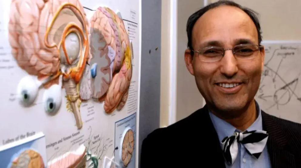 A man with glasses and bow tie stands next to a wall display of the human brain