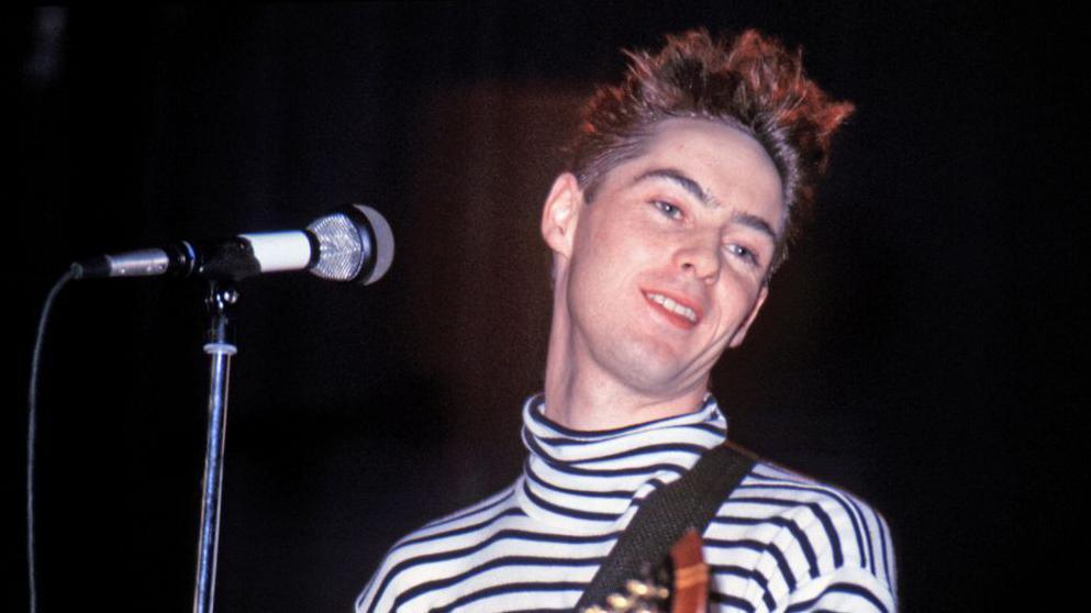 Roddy Frame performing on stage in the eighties. He is wearing a black and white polo top. He is in front of mic and has a guitar over his shoulder 