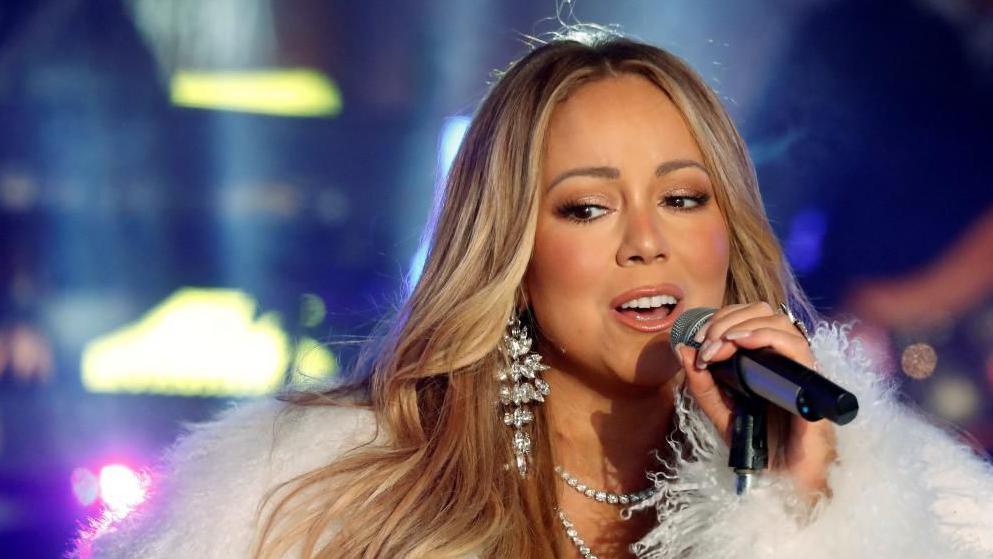 Mariah Carey singing into a microphone. She is wearing a white fluffy jacket and silver earrings. 