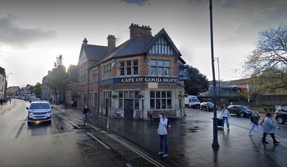 The front of the Cape of Good Hope pub on a damp day. There are some people walking in front. A car is driving by on the street. There is a man riding a bicycle by the pub.