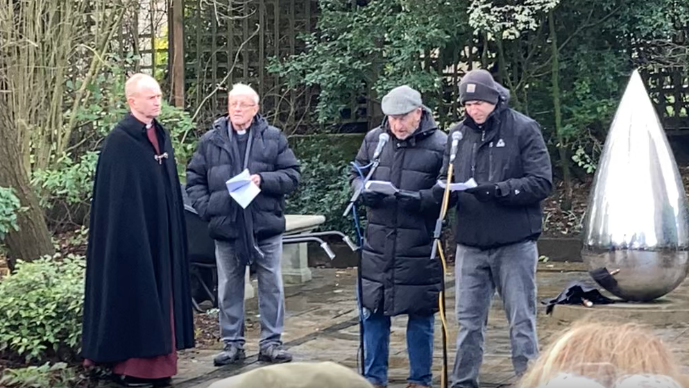 Reverend Canon Matthew Vernon stands and looks on at two other gentleman speaking into microphones during the Holocaust Memorial Day event. They were coats and the two men speaking wears hats as they read pieces of paper in their hands.