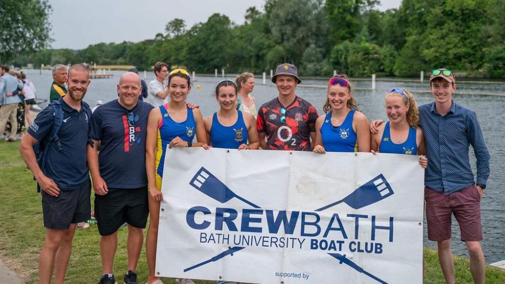 A group of men and women holding up a Crew Bath, Bath University Boat Club sign. Four of the women are in blue rowing kit and four men are in workout kit. They are standing in front of a river. 