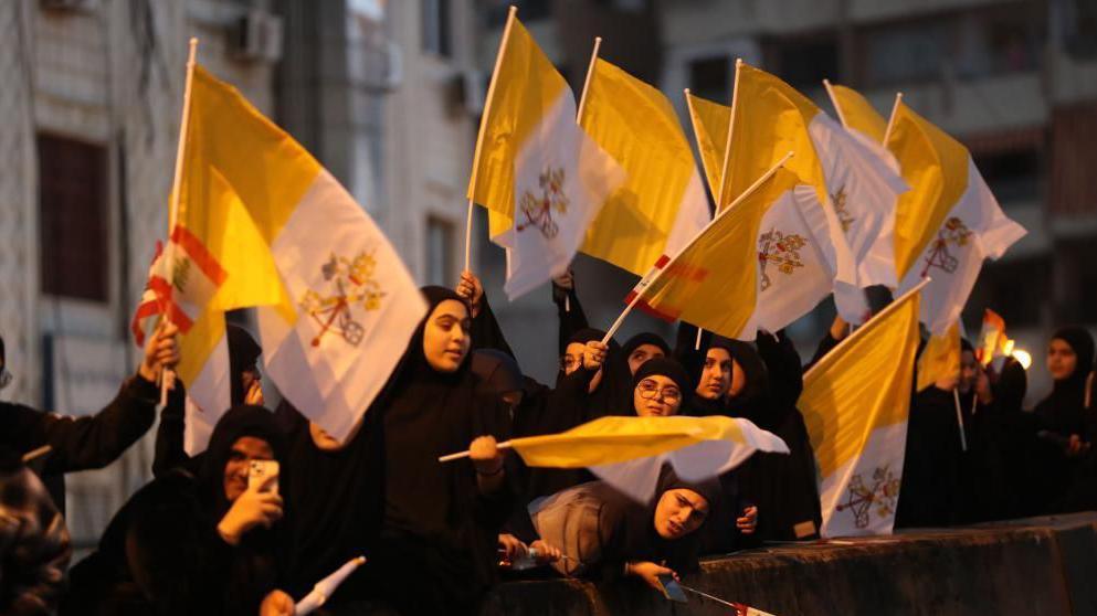 Excited young women in Islamic scarves wave papal flags outside a building.