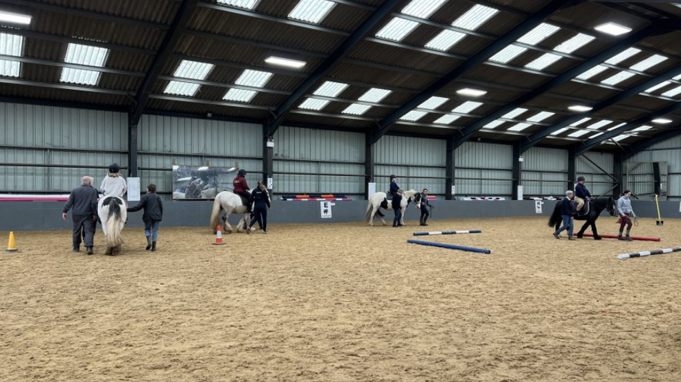 A view of the horse riding school. There are four horses, with four riders on them. The ground is light brown and there are blue and white tubes on the ground. There are people stood either side of the horses, leading them around the school.