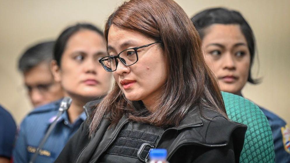 Alice Guo wearing glasses and a black jacket is seated at a table during one of the senate hearings in Manila. Begind her two female officers are visible in their navy uniforms.   