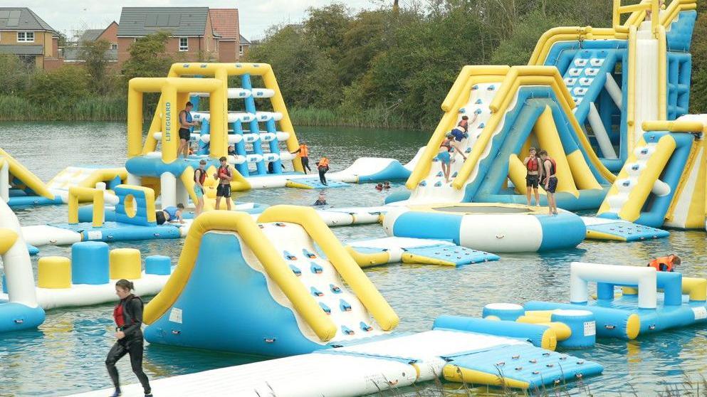 A view of an aqua park has lots of blue, white and yellow inflatables to play on and try to get across on an expanse of water. A number of people trying the course are seen wearing swimming suits, trunks and wet suits and there is a man standing on an inflatable bearing the word "lifeguard"