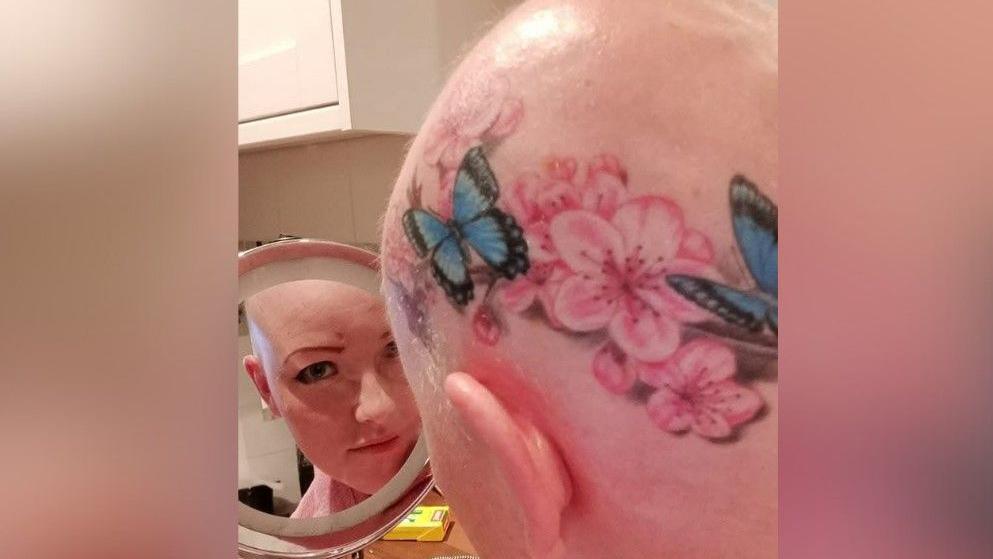 The back of Ariane Murrell's head as it shows off her large tattoo, which has pink blossoms and blue and black butterflies. She is looking at the camera via a handheld mirror.