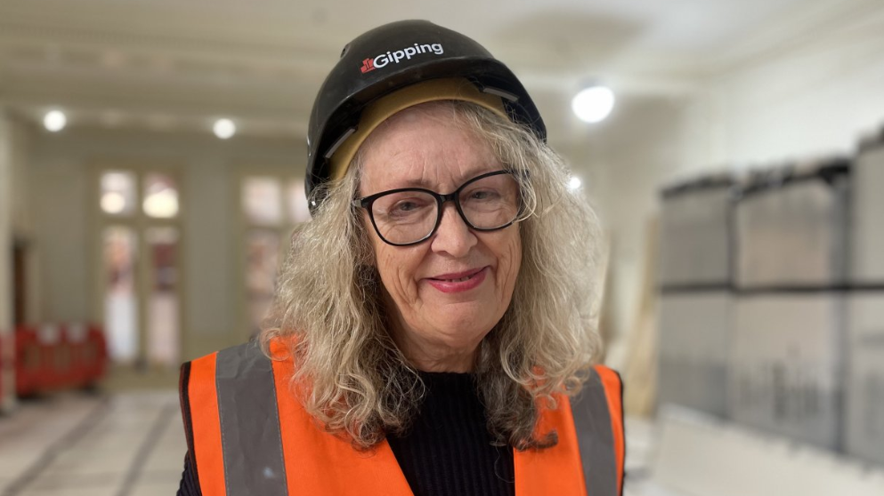 Woman with long blonde/grey hair wearing spectacles, an orange hi-vis vest and a black hard hat with Gipping written across the front