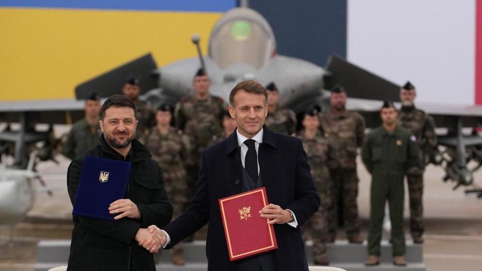 Two men stand smiling in front of a fighter jet in a hangar.