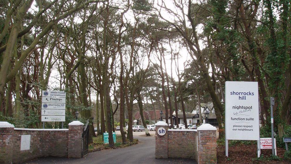 A brick wall entrance with iron gates opens to the former nightclub site. The sign reads Shorrocks Hill nightspot Please respect our neighbours. The area is surrounded by woodland and tall trees. 