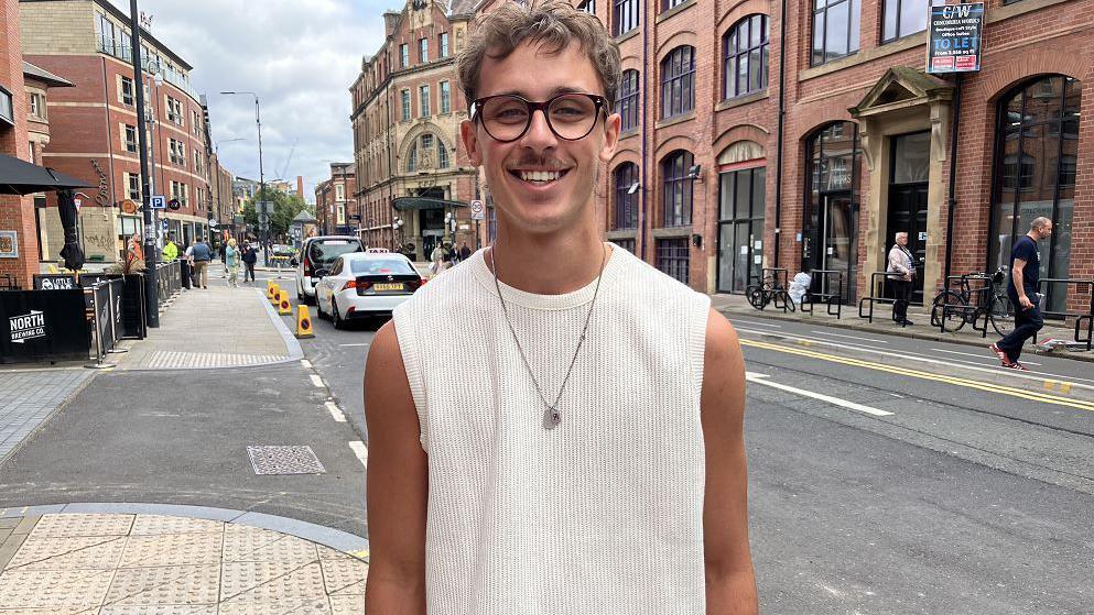 A man wearing glasses and a cream coloured sleeveless top stands in a street in Leeds.