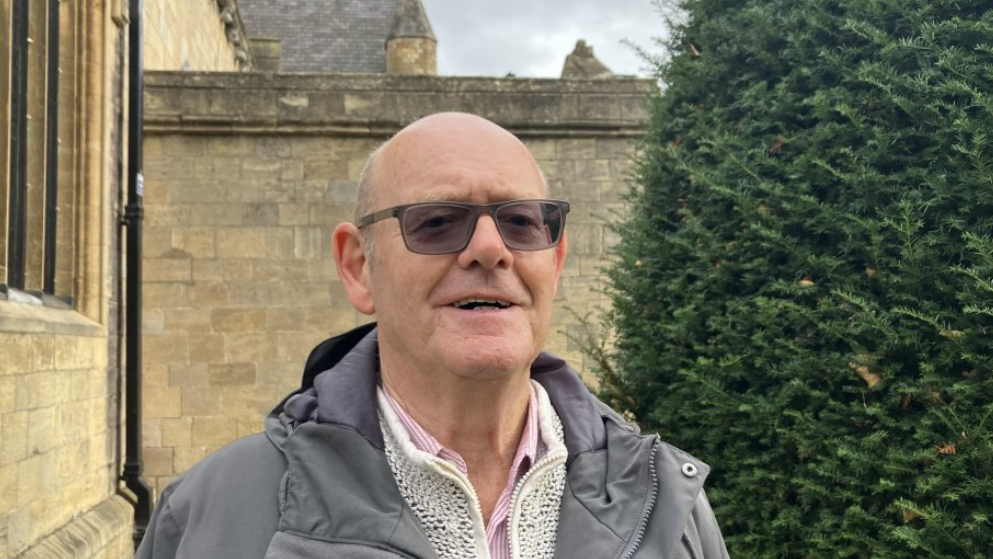 Bald smiling man wearing sunglasses. He is wearing a grey hooded anorak/jacket and cream cardigan with a pink and white striped shirt. He is standing in front of a church and a Yew bush. The sky is cloudy and grey