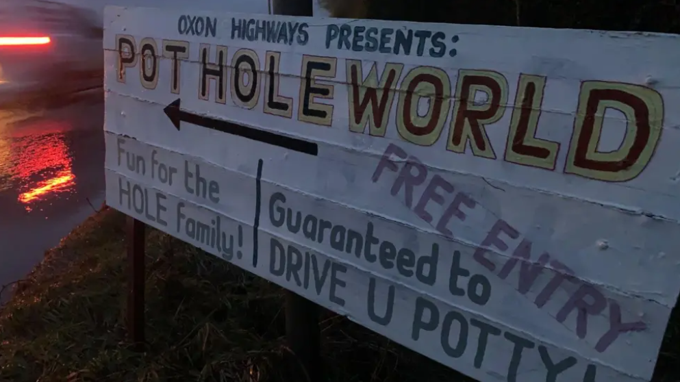 A sign reading: "Oxon Highways presents: Pothole World", "fun for the hole family" and "guaranteed to drive you potty". It is right next to a road.