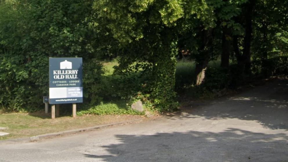 A drive off a main road with Trees and grass alongside the road. A sign reads Killerby Old Hall cottages, lodges, caravan park.