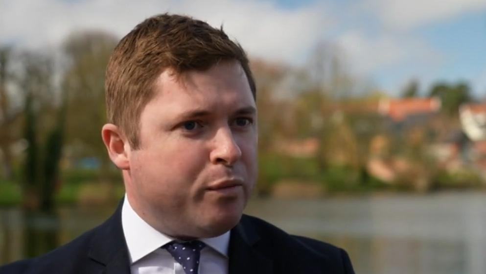 A man wearing a shirt and tie, and a black jacket. He has short light brown, slightly red hair, and is clean shaven. He is near a lake, and there are buildings in the backdrop, in soft focus.