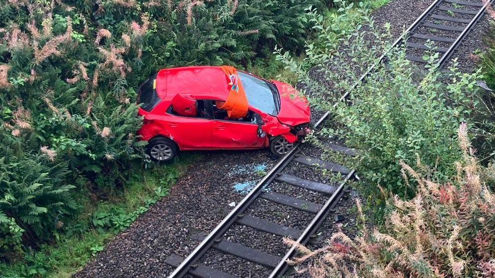 A badly-damaged small red car sits on a railway line surrounded by vegetation