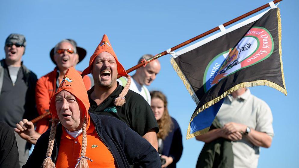 Competitors and spectators wearing hats and carrying a flag at 2013's World Stone Skimming Championships