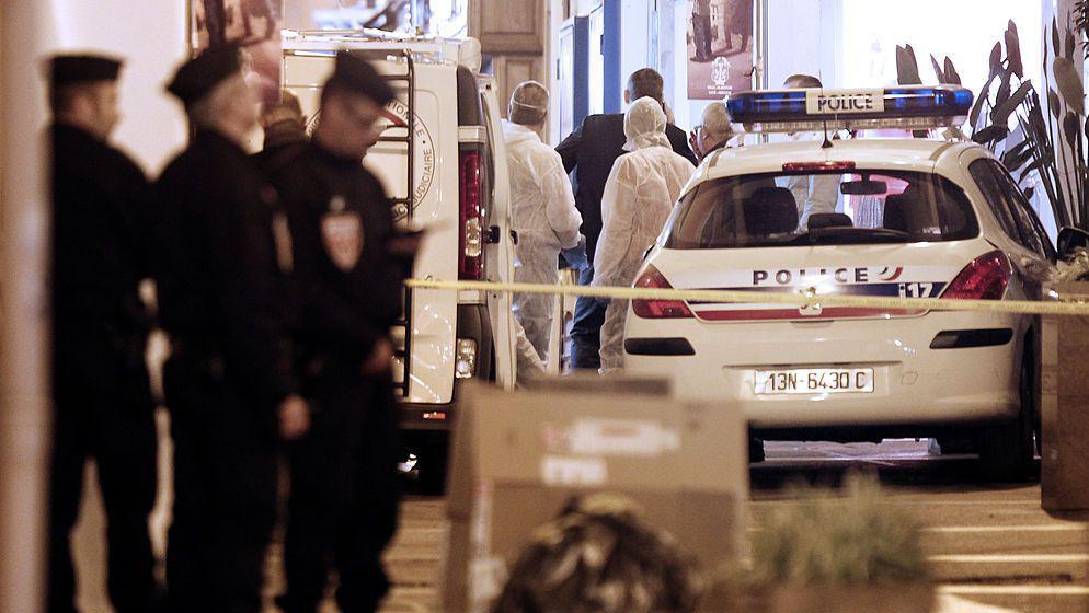 French policemen and forensics investigate around a crime scene - with police in dark uniforms unfocused in the foreground and a white police car and forensics officers in white suits in the background