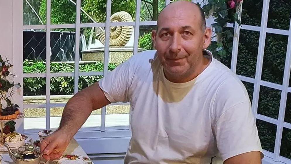 A man in his 40s with a shaved head and white T-shirt, holds a teapot while sitting at a table. A garden is visible through the window behind him