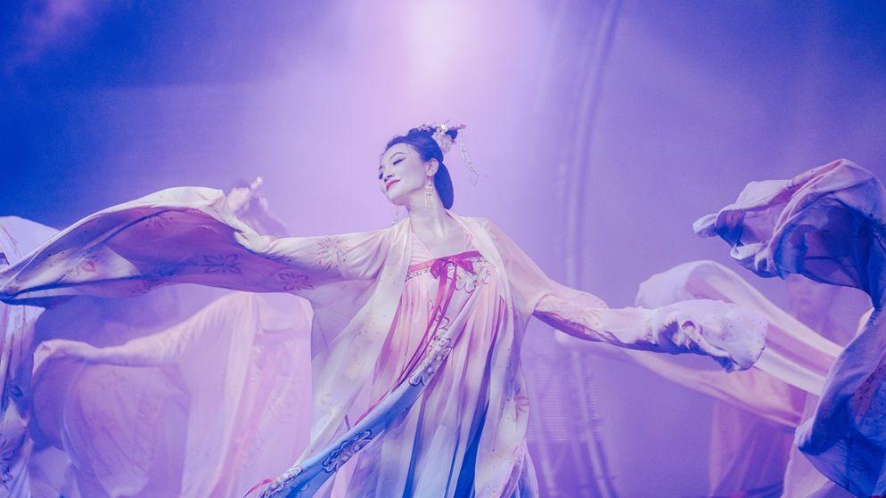 An event during Lunar Chinese New Year. A Chinese woman with her hair tied back tidily at her neck is on stage. She has sharp, contrasting make-up on. The stage lighting is pink and purple. She wears flowy, light-pink dress and appears to be in a group dance arrangement. She has a serene expression.