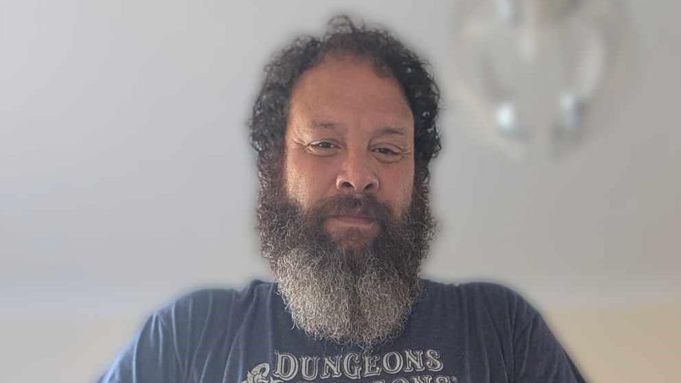 A  man with dark curly hair and a large bear looks down at the camera and smiles, he is wearing a blue T-shirt with the word "Dungeons" on it. 