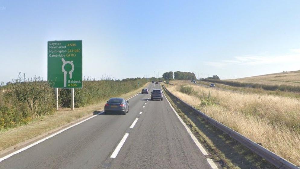 Cars travel along a dual carriageway with long grass and fields either side of the road, and a sign indicating an upcoming roundabout.