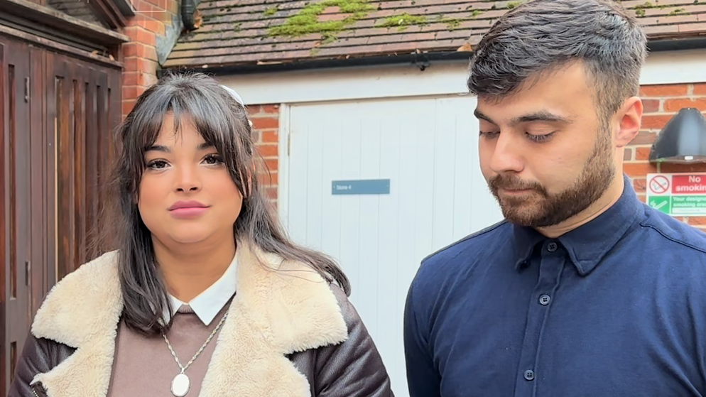 The parents of Artemis Sanson, Storm and Jamie, stand outside, posing for photographers. She wears a brown bomber jacket and brown top with a silver pendant, and has shoulder length brown hair. He wears a dark blue shirt buttoned up to the nexk with no tie, and has dark hair and a trimmed beard.