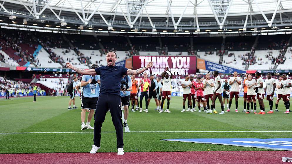 Manager Nathan Jones raises both hands to celebrate in front of his players while wearing a dark blue outfit and white trainers as Charlton Athletic Football Club head coach after their win against West Ham United at the London Stadium