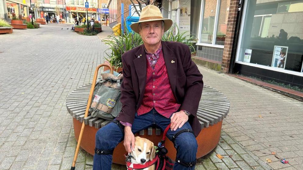 A man in a Purple blazer, red waistcoat, jeans and a beige hat sits on a circular bench in a town centre while petting his brown and white dog. A curved walking stock and a grey bag are next to them.  