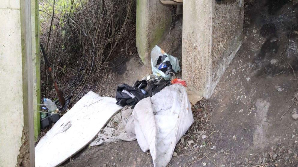 A makeshift camp under a small bridge in Brantham. A white crumpled up duvet can be seen alongside a bag of rubbish and other items stress across the ground
