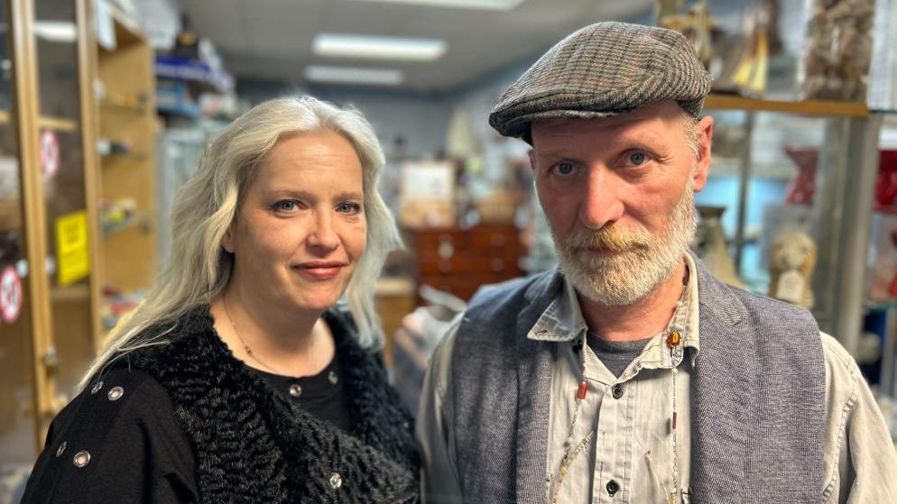 A woman with long white hair wearing black and a man in a flat cap, with a white beard and a waistcoat, stand in an antique shop. 