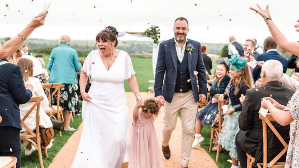 A happy bride and groom walk down the aisle holding the hand of a young bridesmaid in a pink dress.