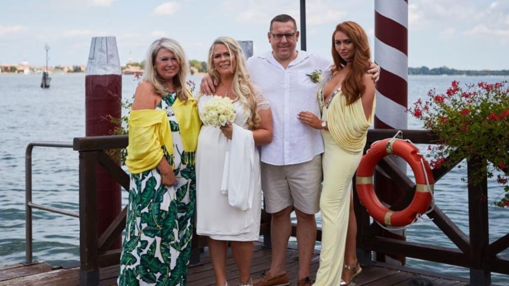 Samantha Southern is wearing a white dress and holding a bouquet of flowers. She is standing next to her husband Glen who is wearing a white shirt and beige shorts. They are standing next to two other family members in front of the sea.