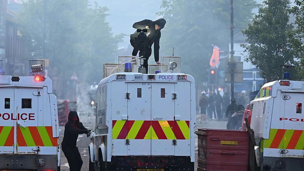 Three police vans in Northern Ireland lined up - a man in all black with his hood up crouches on the roof of the middle van while another hooded man stands next to it on the ground. There are crowds standing around in the background and the sky is smoky.