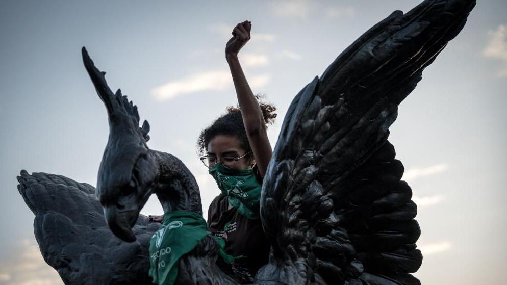 A woman who is covering her mouth with a green bandanna lifts her left fist in a gesture of defiance as she is perched on top of a statue during a protest in Lima.