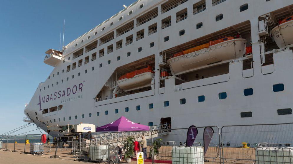 A large cruise ship berthed at Invergordon. The boat is white and several storeys high. The word "Ambassador" is in purple on the side of the ship.