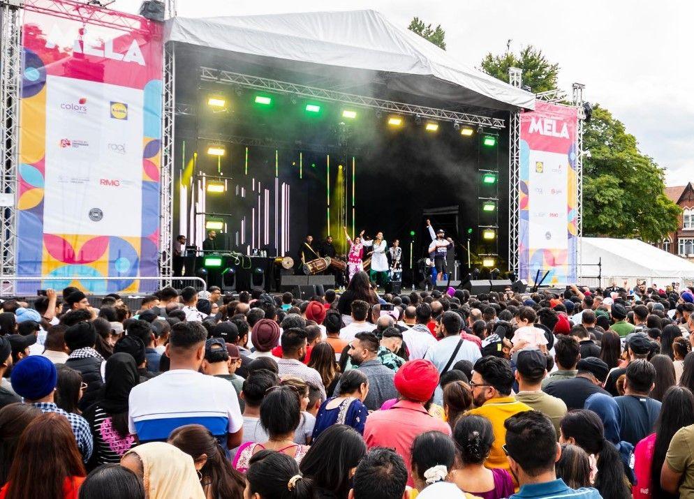 A crowd watching a musical performance on a large outdoor stage with sponsorship banners either side