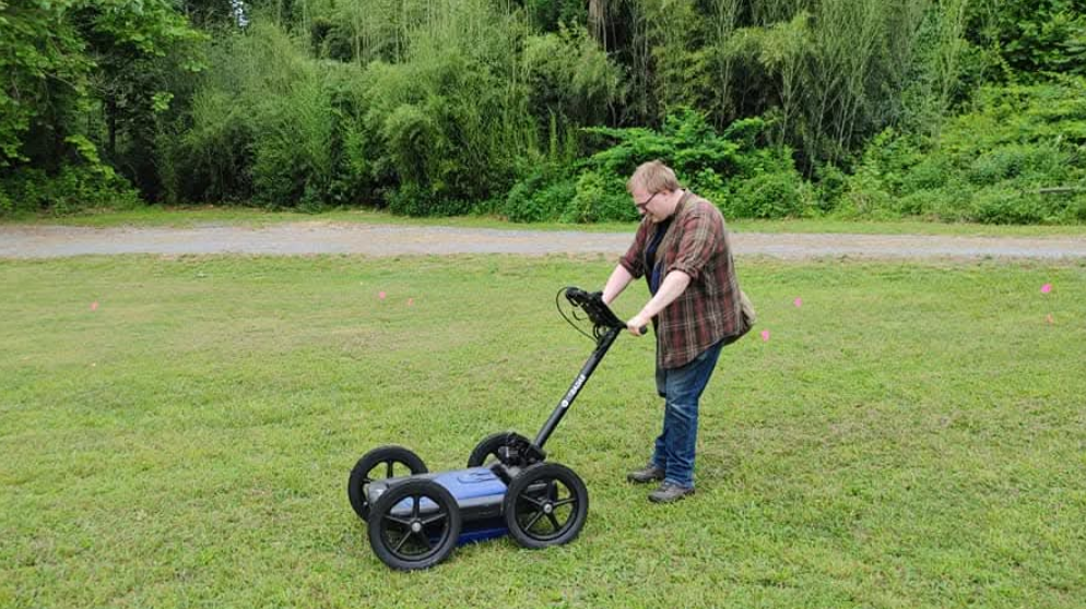 A man with fair hair and glasses and wearing a checked-shirt and jeans pushes a four-wheeled machine along a field of grass