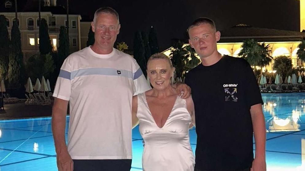 A man, woman and teenager in front of a swimming pool while on holiday - the man is wearing a white shirt and shorts, the woman a white dress and the teenager a black shirt and shorts