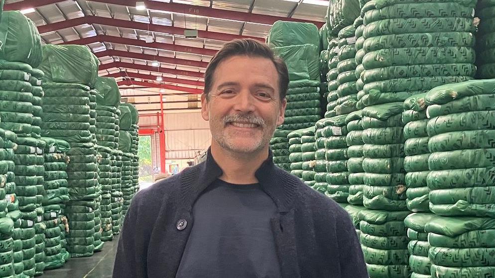 A man with short brown hair and a grey moustache stands in a warehouse surrounded by green sacks on wool fleeces. He is wearing a navy cotton T short and navy wool cardigan